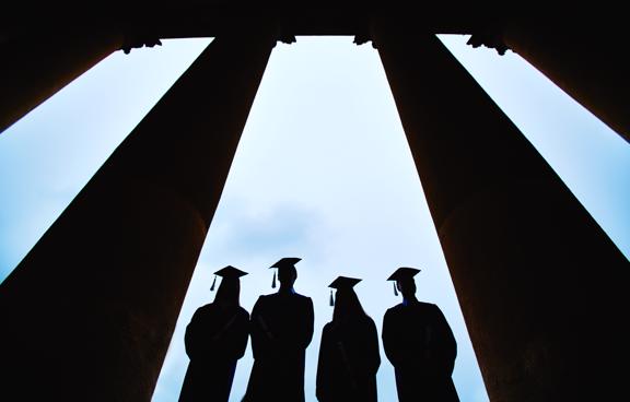 Photo of four graduates shown in dark figure with clouds behind them and columns to each side.
