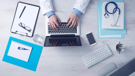 Stock image of a doctor shown with glasses, laptop, and notes.